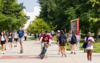 Students walk, bike, and scooter along a tree-lined campus path on the first day of classes, with a wayfinding sign pointing toward Bush Theatre, the Music Building, and the Library