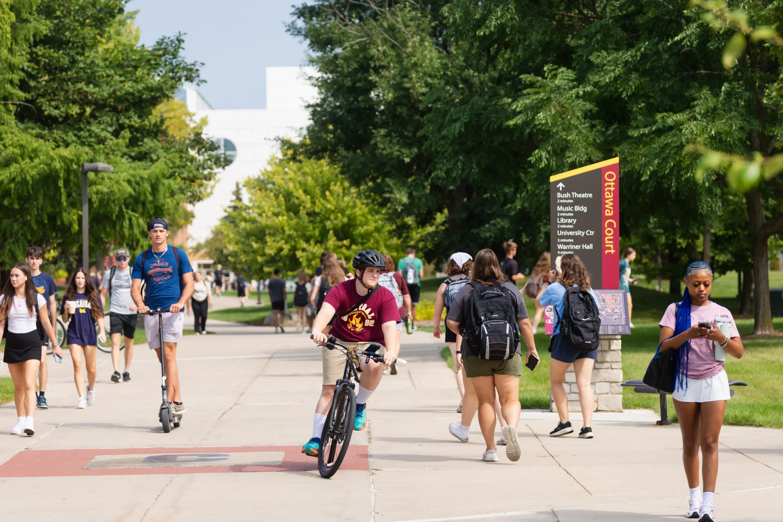 Students walk, bike, and scooter along a tree-lined campus path on the first day of classes, with a wayfinding sign pointing toward Bush Theatre, the Music Building, and the Library